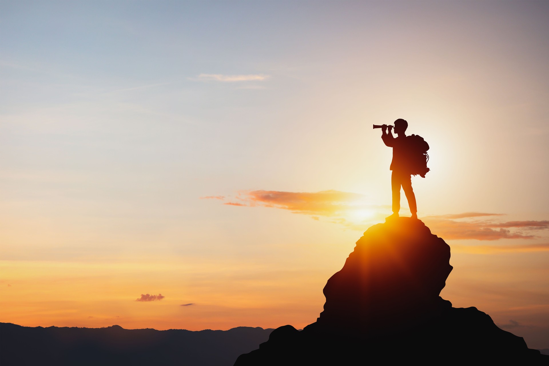 Silhouette of man holding binoculars on mountain peak against bright sunlight sky background.